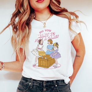 K&ouml;nnte beinhalten: Wei&szlig;es T-Shirt mit einem Motiv von drei M&auml;dchen, die ein Spiel mit einem Ouija-Brett spielen. Der Text auf dem Shirt lautet "Do you want to play a game?"