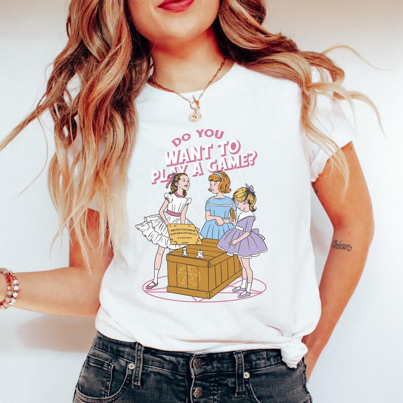 K&ouml;nnte beinhalten: Wei&szlig;es T-Shirt mit einem Motiv von drei M&auml;dchen, die ein Spiel mit einem Ouija-Brett spielen. Der Text auf dem Shirt lautet "Do you want to play a game?"
