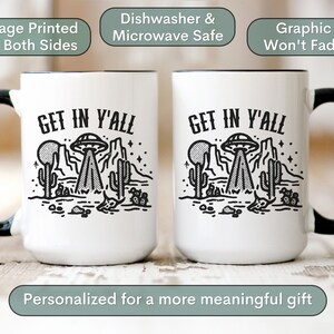 May include: Two white ceramic mugs with black handles and black rims. Both mugs feature a black and white graphic of a UFO over a desert landscape with the text "GET IN Y'ALL".
