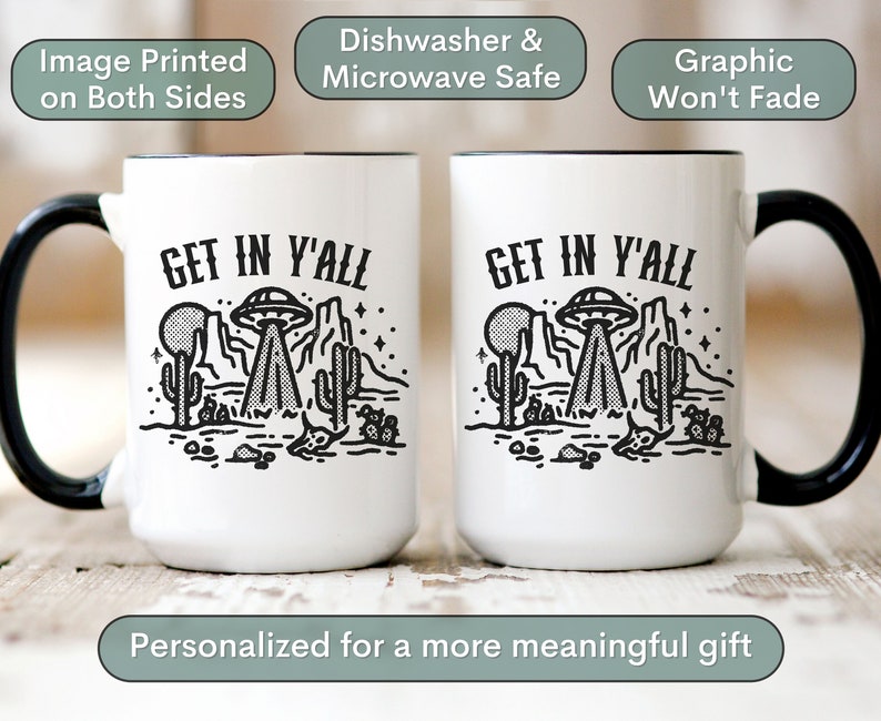 May include: Two white ceramic mugs with black handles and black rims. Both mugs feature a black and white graphic of a UFO over a desert landscape with the text "GET IN Y'ALL".