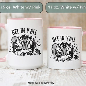 May include: Two white ceramic mugs with pink rims. Both mugs feature a black and white illustration of a UFO over a desert landscape with the text "GET IN Y'ALL". The mug on the left is 15 ounces and the mug on the right is 11 ounces.