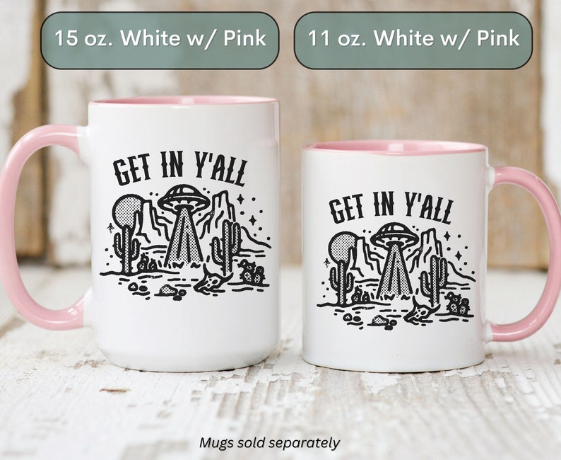 May include: Two white ceramic mugs with pink rims. Both mugs feature a black and white illustration of a UFO over a desert landscape with the text "GET IN Y'ALL". The mug on the left is 15 ounces and the mug on the right is 11 ounces.