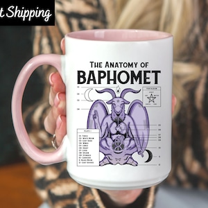 May include: White ceramic mug with a pink handle and interior. The mug features a detailed illustration of Baphomet with anatomical labels and the text "THE ANATOMY OF BAPHOMET". The design includes a pentagram and moon symbols.