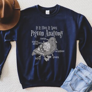 May include: Navy blue sweatshirt with the text "If It Flies It Spies Pigeon Anatomy." The graphic depicts a pigeon with labeled parts like "camera," "microphone," and "battery." A brown hat and blue jeans are partially visible.
