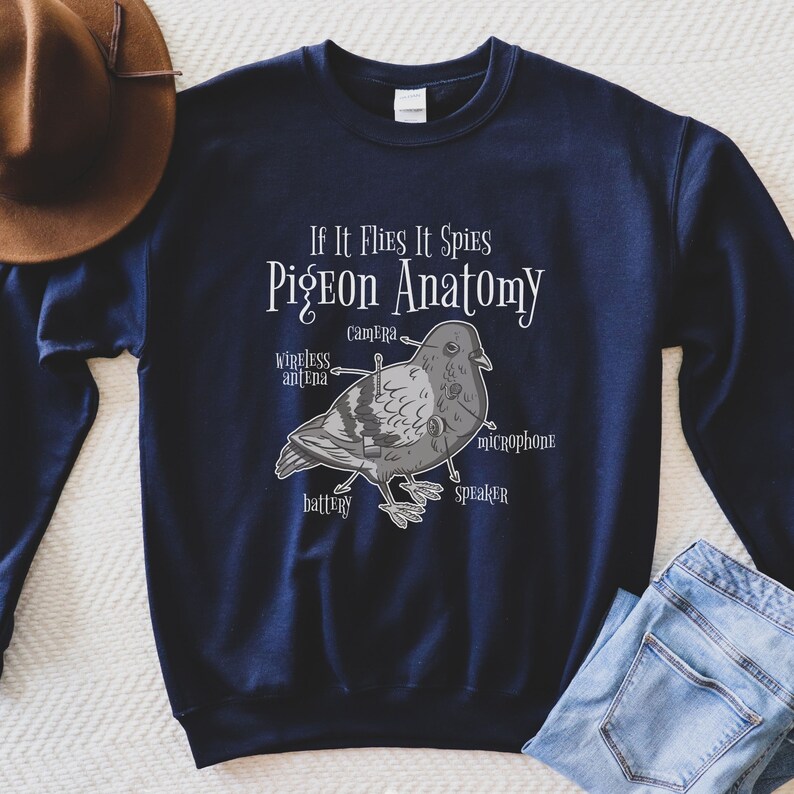 May include: Navy blue sweatshirt with the text "If It Flies It Spies Pigeon Anatomy." The graphic depicts a pigeon with labeled parts like "camera," "microphone," and "battery." A brown hat and blue jeans are partially visible.