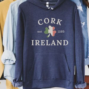 Op de afbeelding: Marineblauwe hoodie met een voorzak en een witte opdruk met de tekst "CORK IRELAND" met een klaverblad in de kleuren van de Ierse vlag. De tekst "est 1185" is ook inbegrepen. De hoodie hangt met andere kleding.