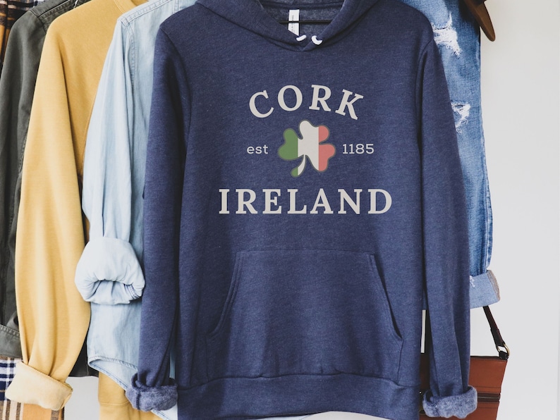 Op de afbeelding: Marineblauwe hoodie met een voorzak en een witte opdruk met de tekst "CORK IRELAND" met een klaverblad in de kleuren van de Ierse vlag. De tekst "est 1185" is ook inbegrepen. De hoodie hangt met andere kleding.