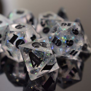 May include: A collection of clear, glitter-filled dice with black numerals. The dice are in various shapes, such as a D20, D6, and D4, and are intended for tabletop role-playing games. The dice are presented on a reflective surface.