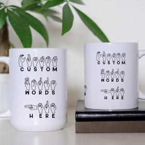 May include: Two white ceramic mugs with black text and images. The mugs feature the text "Custom Words Here" and images of hands signing the letters of the alphabet.