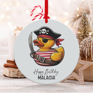 May include: A circular Christmas decoration with a cartoon pirate duck design. The duck wears a black hat, eye patch, and a red and white striped top, holding a sword. The text "Happy Birthday MALACHI" is written on the ornament.