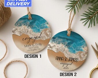 Könnte beinhalten: Zwei runde Ornamente mit Strandmotiv, mit blau-weißem Wellendesign. Eines zeigt "engaged" mit Namen und Datum, das andere "engaged" mit Namen und dem Jahr 2025. Beide haben eine Aufhängeschnur.