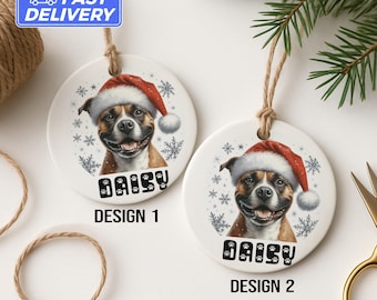 May include: Two round ceramic Christmas ornaments featuring a smiling dog wearing a Santa hat. The dog's name, "Daisy", is printed below the image. Snowflake and paw print designs surround the dog. The ornaments are hanging by twine.