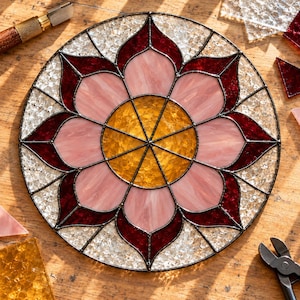 May include: A circular stained glass panel featuring a central yellow sunburst, surrounded by light pink petals and deep red outer petals, all encased in a dark metal frame with clear textured glass accents in the outer border.