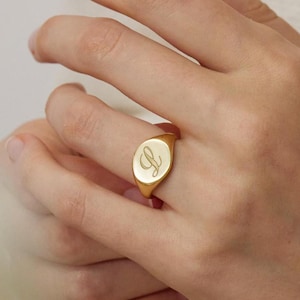 Puede incluir: Un anillo de sello dorado con una cara ovalada, con una inicial en escritura cursiva. El anillo se lleva en un dedo, mostrando su superficie pulida. El diseño del anillo es simple y elegante.