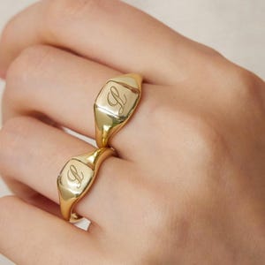 以下が含まれることがあります： 小さな彫刻が施された2つの金のシグネットリングが手に着用されています。リングは磨かれた仕上げで、クラシックでエレガントなデザインです。小さなペンダントが付いた金のネックレスも表示されています。