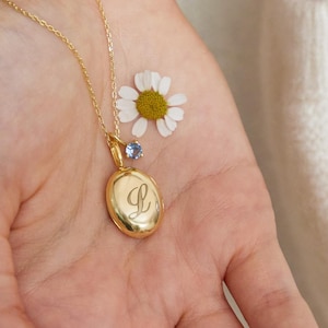 Puede incluir: Un collar con un colgante ovalado dorado con la letra "L" grabada. El colgante cuelga de una delicada cadena. Una pequeña gema azul y una margarita blanca están junto al colgante. El fondo es un suéter blanco.