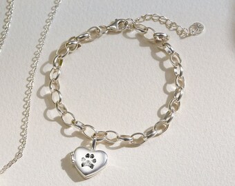 Puede incluir: Un conjunto de collar y pulsera de plata con colgantes en forma de corazón que presentan un diseño de huella de pata. Los colgantes son abatibles y se pueden usar para guardar un pequeño recuerdo.