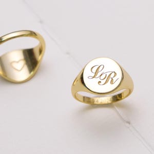 May include: Two gold signet rings. One ring features a flat circular face engraved with the cursive letters LR. The second ring is positioned to show the interior of the band, which is engraved with a small, simple heart outline symbol.