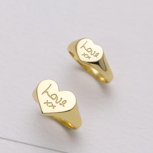 Puede incluir: Dos anillos dorados con forma de corazón. La palabra "love" está inscrita en cada corazón, junto con dos símbolos "xx". Los anillos están sobre un fondo blanco.