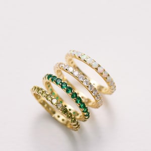 May include: Four gold rings with different colored gemstones. The rings are stacked on top of each other. The top ring has white gemstones, the second ring has green gemstones, the third ring has green gemstones, and the bottom ring has yellow gemstones.