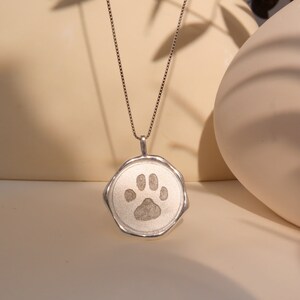 May include: A silver necklace with a round pendant featuring a paw print design. The pendant has a textured, slightly irregular edge.