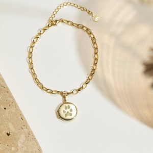 May include: A gold chain bracelet with a small round charm featuring a paw print. The charm is attached to the chain with a small loop.