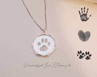 May include: A silver necklace with a round pendant featuring a paw print. The pendant is on a white surface with black ink paw prints, a heart, and a handprint.