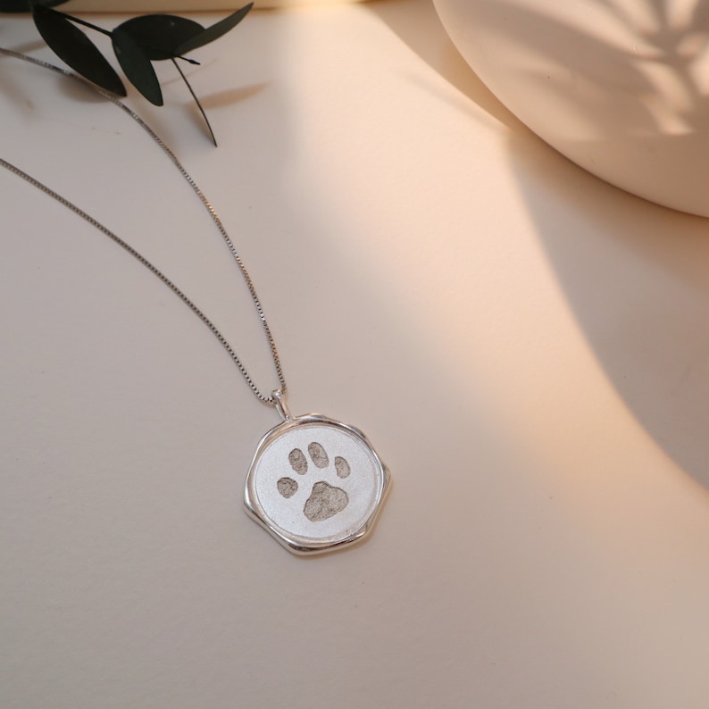 May include: A silver necklace with a round pendant featuring a paw print design. The pendant has a textured, slightly irregular edge.