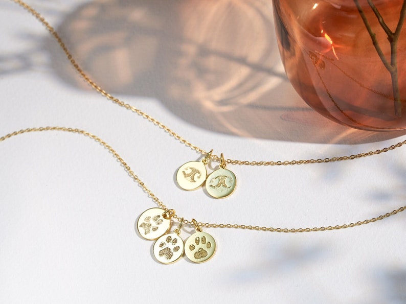 May include: A gold necklace with three charms: two round charms with initials and one charm with a paw print. The necklace is on a white surface.