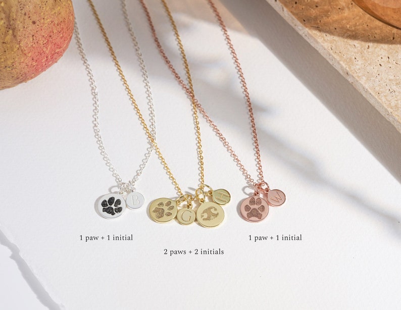 May include: Three silver necklaces with charms. The first necklace has a single paw print charm and a single initial charm. The second necklace has two paw print charms and two initial charms. The third necklace has a single paw print charm and a single initial charm.