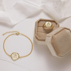 Peut inclure: Un bracelet en cha&icirc;ne dor&eacute;e avec un pendentif rond grav&eacute; et une bague signet dor&eacute;e dans un &eacute;crin &agrave; bijoux en velours beige.