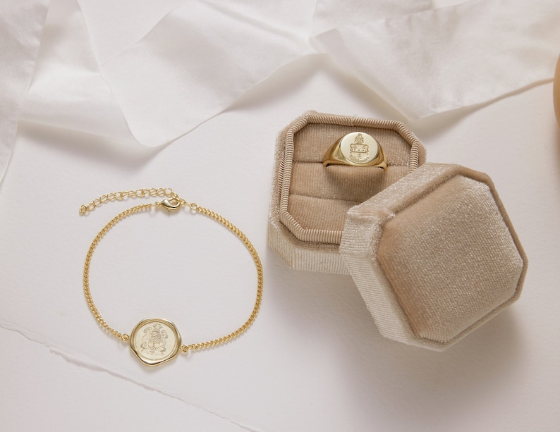 Peut inclure: Un bracelet en cha&icirc;ne dor&eacute;e avec un pendentif rond grav&eacute; et une bague signet dor&eacute;e dans un &eacute;crin &agrave; bijoux en velours beige.