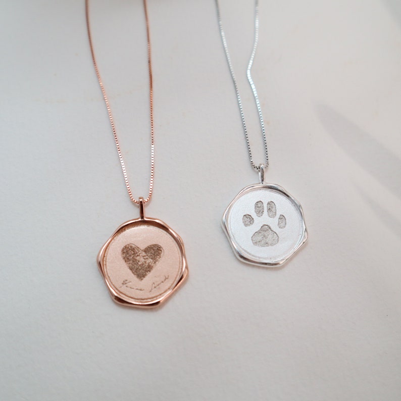 May include: Two necklaces with pendants featuring engraved paw prints and a heart. The silver necklace has a paw print pendant, and the rose gold necklace has a heart pendant with the words "true friend" engraved on it.