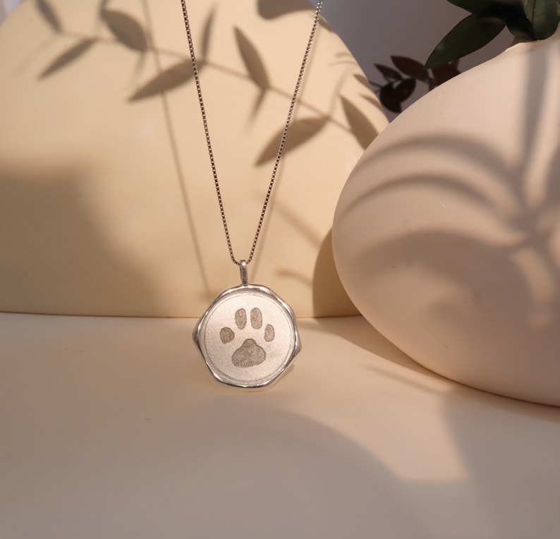 May include: A silver necklace with a round pendant featuring a paw print design. The pendant has a textured, slightly irregular edge.