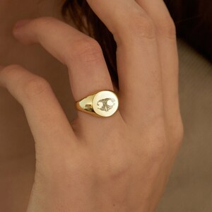 May include: A gold signet ring with a detailed design on the top. The ring is oval shaped and has a smooth, polished finish.