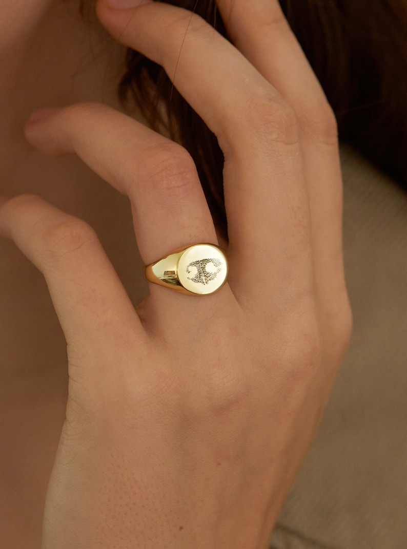 May include: A gold signet ring with a detailed design on the top. The ring is oval shaped and has a smooth, polished finish.
