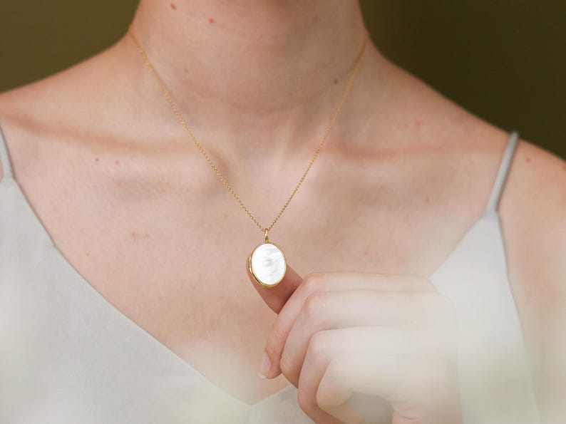 Op de afbeelding: Een gouden ketting met een witte ovale hanger.