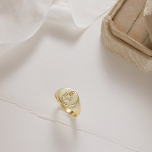 Peut inclure: Une bague signet en or avec un motif grav&eacute; d&eacute;taill&eacute;. La bague est sur une surface blanche.