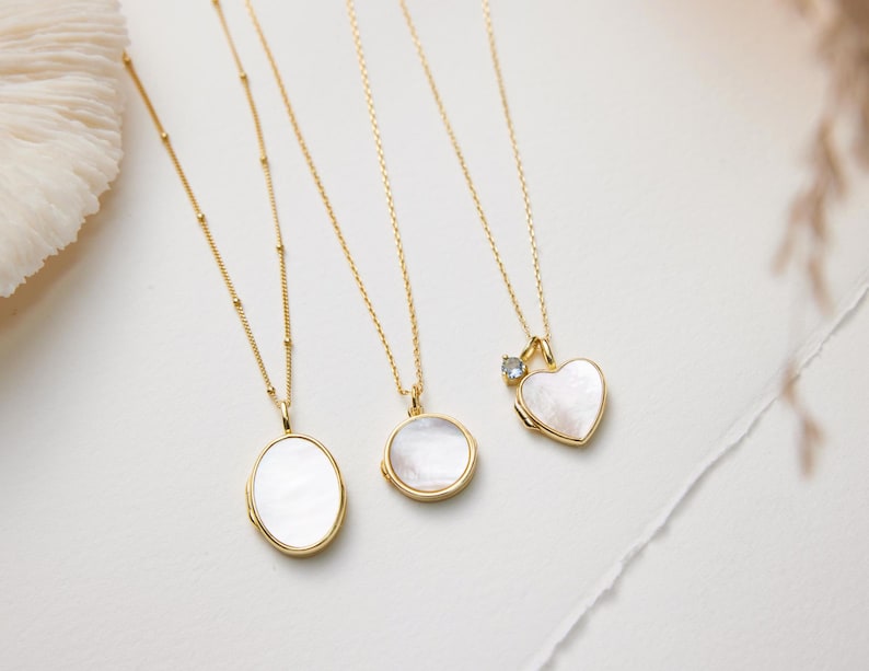 Op de afbeelding: Vier gouden kettingen met verschillende hanger stijlen. Elke hanger is gemaakt van wit parelmoer en heeft een andere vorm: ovaal, rond, hart en hart met een kleine diamant.
