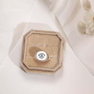 May include: A silver signet ring with a white stone and a detailed design on the top. The ring is displayed in a beige velvet ring box.