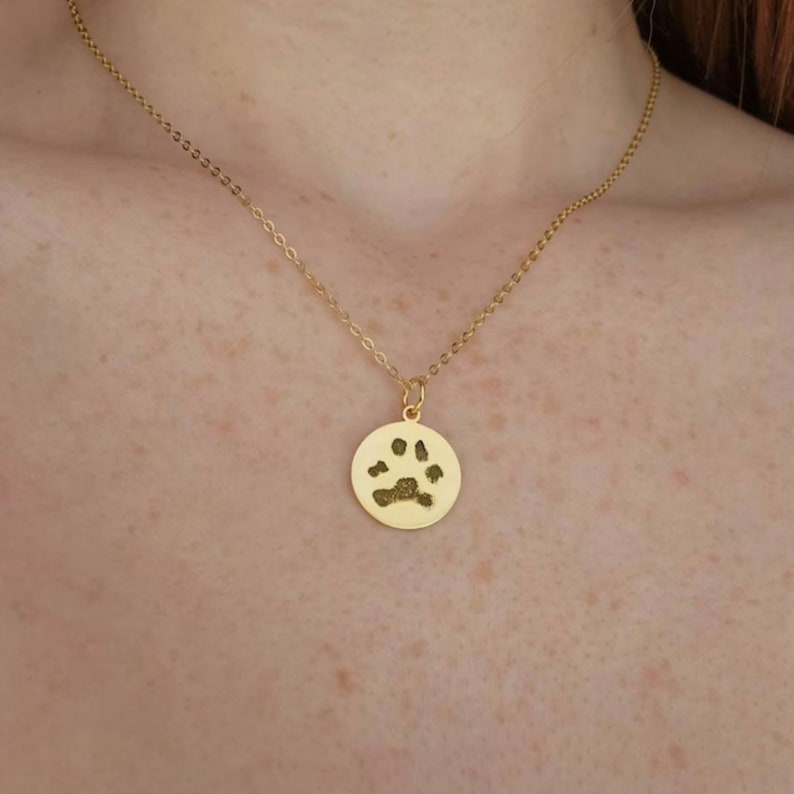May include: A gold necklace with a round pendant featuring a paw print. The pendant is a memorial piece for a pet.