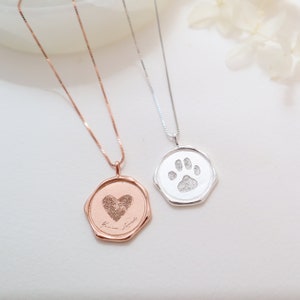 May include: Two silver necklaces with octagonal pendants. One pendant features a heart with a paw print inside, the other features a paw print. Both pendants have a handwritten inscription.