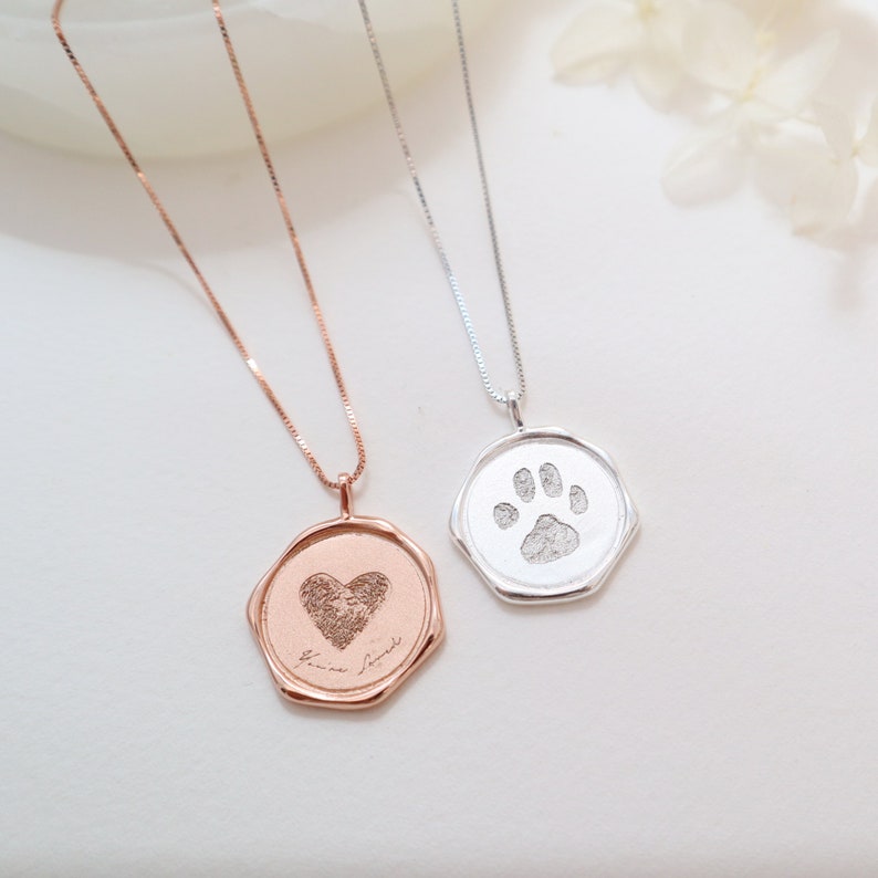 May include: Two silver necklaces with octagonal pendants. One pendant features a heart with a paw print inside, the other features a paw print. Both pendants have a handwritten inscription.
