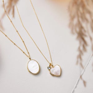 Op de afbeelding: Twee gouden kettingen met delicate kettingen en hangers. De ene hanger is een wit ovaal medaillon, de andere is een wit hartvormig medaillon met een kleine heldere steen.