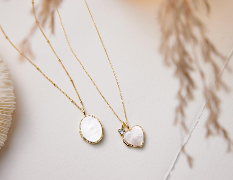 Op de afbeelding: Twee gouden kettingen met delicate kettingen en hangers. De ene hanger is een wit ovaal medaillon, de andere is een wit hartvormig medaillon met een kleine heldere steen.