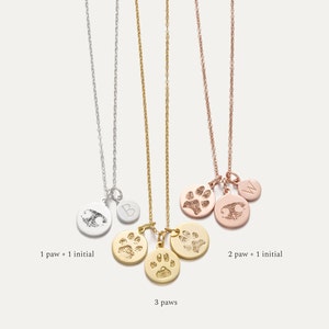 May include: Three gold necklaces with paw print charms. The first necklace has one paw print charm and one initial charm. The second necklace has two paw print charms and one initial charm. The third necklace has three paw print charms. The necklaces are on a white background.