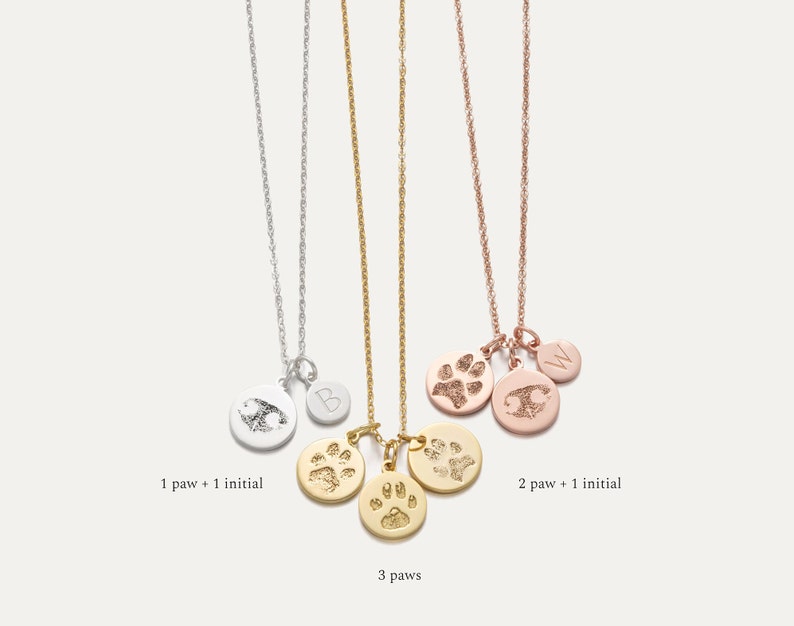May include: Three gold necklaces with paw print charms. The first necklace has one paw print charm and one initial charm. The second necklace has two paw print charms and one initial charm. The third necklace has three paw print charms. The necklaces are on a white background.