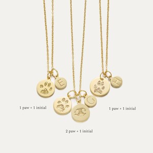 May include: Three gold necklaces with charms. The charms are round and feature a paw print and an initial. The necklaces are on a white background. 1 paw + 1 initial, 2 paw + 1 initial, 1 paw + 1 initial.