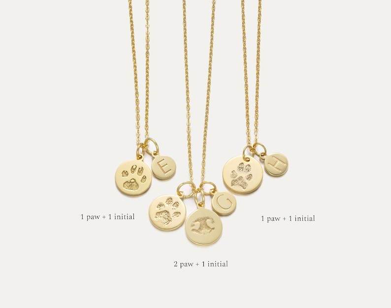 May include: Three gold necklaces with charms. The charms are round and feature a paw print and an initial. The necklaces are on a white background. 1 paw + 1 initial, 2 paw + 1 initial, 1 paw + 1 initial.