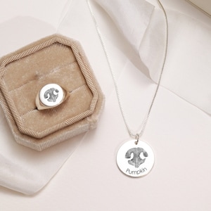 May include: A silver necklace with a round pendant featuring a black and white paw print. The pendant is engraved with the word "Pumpkin". A silver ring with a round top featuring a black and white paw print is in a brown velvet ring box.